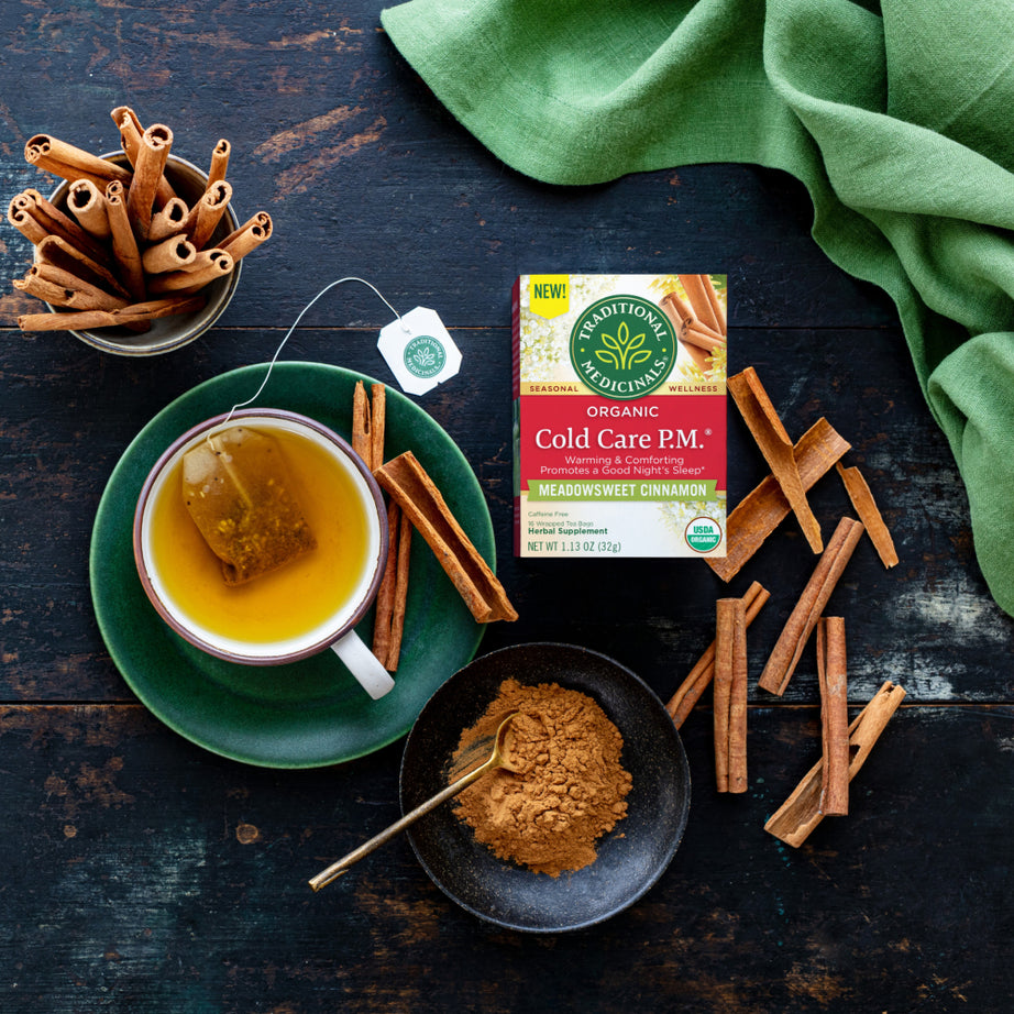 A cup of tea with cinnamon sticks and powder is placed on a dark table, next to a box of Cold Care P.M.® Tea, an organic nighttime blend that promotes a good night's sleep. View 2