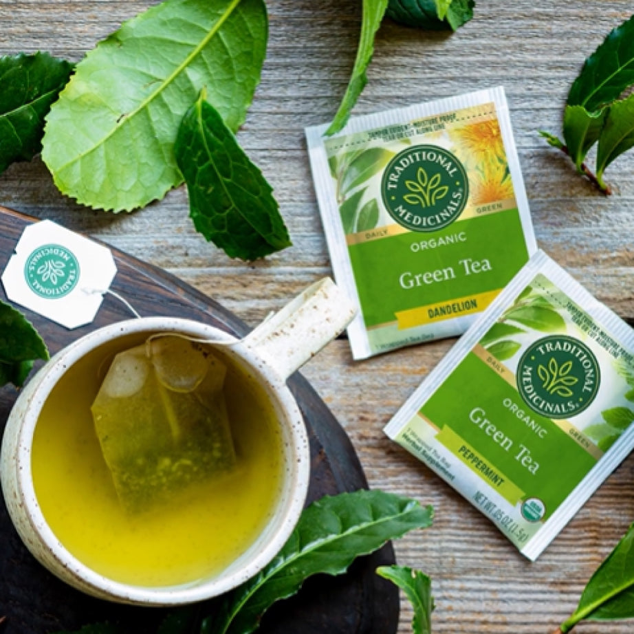 A cup of Green Tea Peppermint with tea bags sits on a wooden surface, surrounded by fresh green leaves. View 2