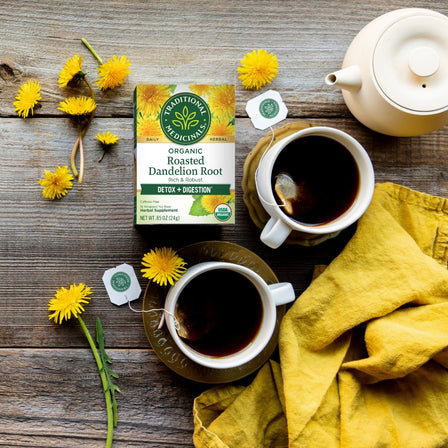 2 cups and 1 teapoy filled with hot tea, a box of Roasted Dandelion Root Tea, and fresh  dandelion root offers a refreshing boost for healthy digestion.